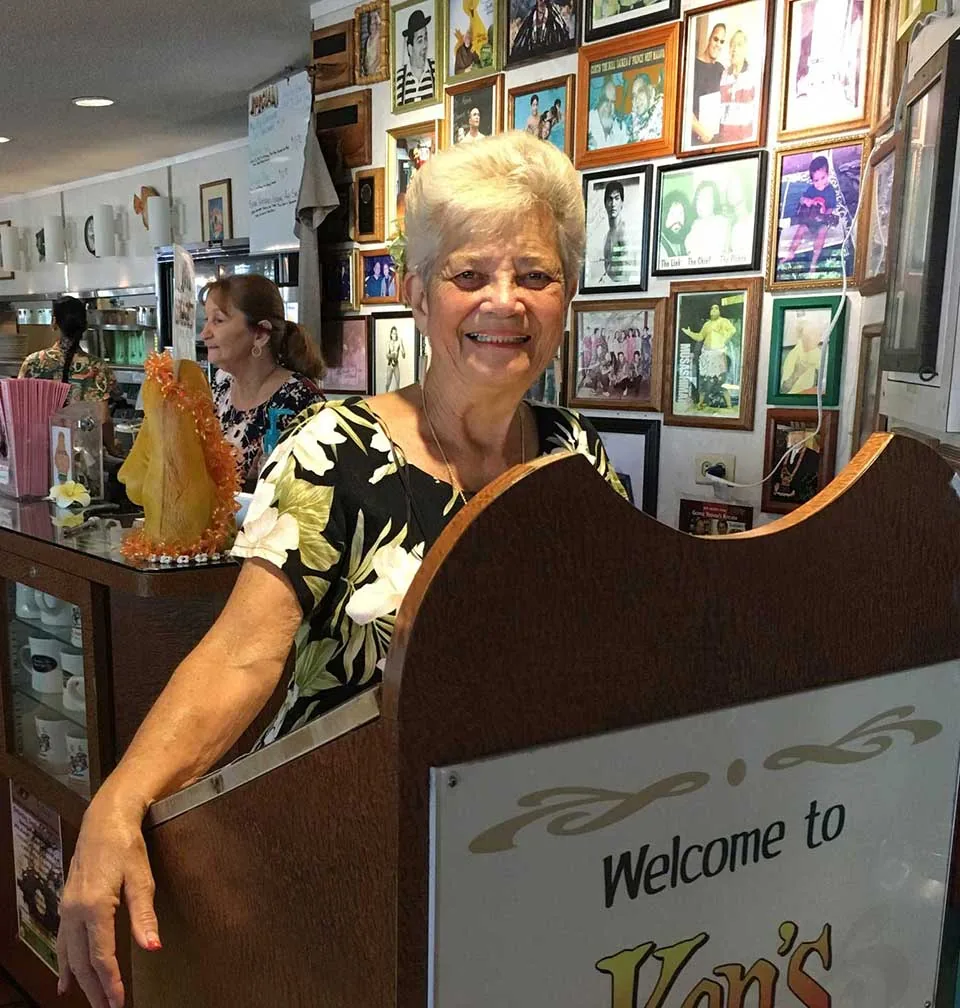 A smiling greeter at the Welcome to Ken's host stand, in front of a wall packed with framed photos of guests and visitors collected over the years