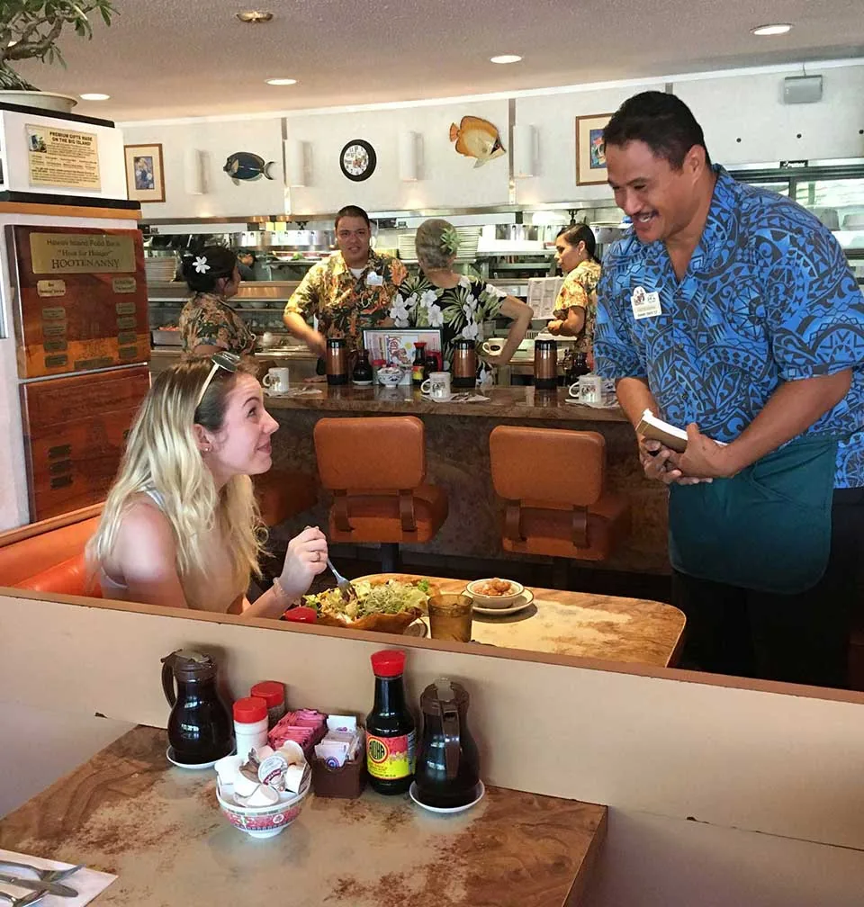 A Ken's server in an aloha shirt taking an order from a guest in a diner booth, with counter stools, syrup caddies on the table, and the open kitchen line behind